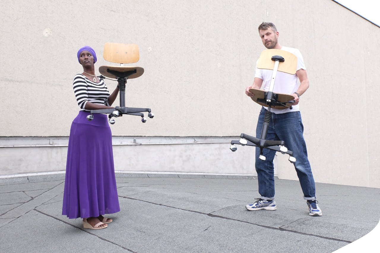 Fatuma Musa Afrah und Jon Hoekstra auf einem Häuserdach, die beide jeweils einen Stuhl in den Händen halten