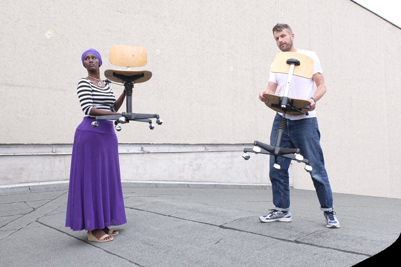 Fatuma Musa Afrah und Jon Hoekstra auf einem Häuserdach, die beide jeweils einen Stuhl in den Händen halten
